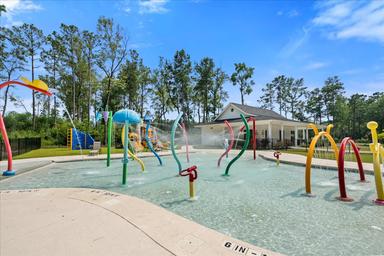Children's splash pad/shallow pool area with colorful water features and clubhouse in background — highlights family-friendly pool amenities located on the campground grounds.