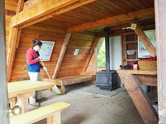Person sweeping the interior of wood cabin with wood table and bunkbed with a black stove