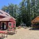 A red building with ice cream cone decoration, tall pine trees in the background, there are picnic tables, a gazebo, and a woodpile shelter in an outdoor area with gravel ground.