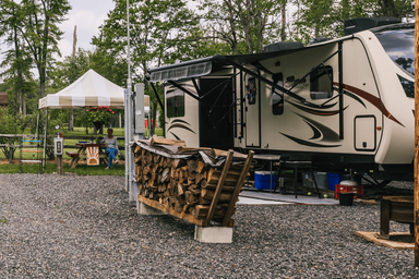 Close view of an RV on a gravel pad with picnic table, firewood stack and utility hookups visible — clearly depicts the RV camping area and site amenities on the property.