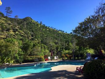 A sunny poolside scene with clear blue water, surrounding trees and hills, lounge chairs, and a bright sky.