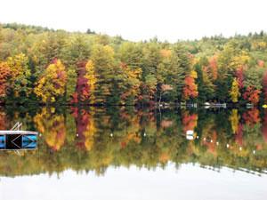 Reflective lakeshore with vivid fall foliage and a swim platform visible at left — another clear view of the beachfront and natural beauty on the property.