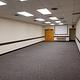 Interior view of the small conference room. Gray carpet flooring with 2 doors at the far end.  Ceiling mounted video screen on far end of room.