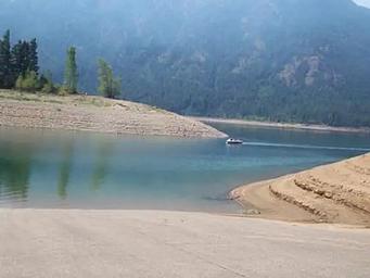 View of boat ramp leading into lake with sloped earthen banks, looking beyond to a small boat and tree covered slopes above high water line.