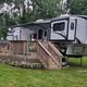 Large camper trailer with wooden deck and stairs in a green outdoor setting.