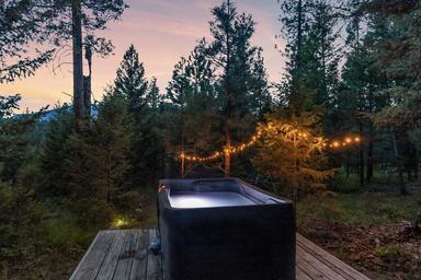 An outdoor hot tub on a wooden deck in a forest, illuminated by string lights hanging from trees at dusk.