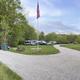 Panoramic view of the campground grounds showing gravel roads, RVs, picnic tables, flagpole and wooded backdrop — good overall site/layout overview (from the home page).