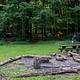 Metal fire ring/grill surrounded by logs and picnic tables in open field.  Multiple tents visible in the distant woods.