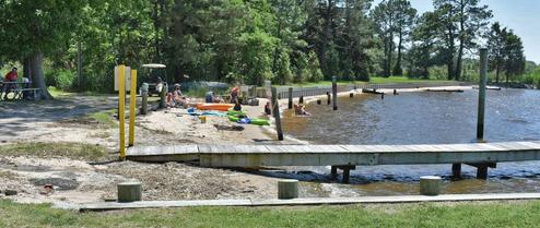 Beach and small dock area with people and waterfront grassy beach — illustrates the campground’s swimming/beach area referenced on the site (Slide-7-Beach).