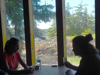 Two people play cards in shadow with view out of Steamer Bay Cabin window visible