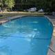 Empty swimming pool in an outdoor setting with a fence, chairs, and trees in the background.