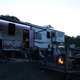 Fifth-wheel RV at a gravel site with family and friends gathered around a campfire at dusk