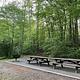 Two wooden picnic tables sit on a gravel pad with a grill on the left and trees behind