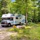 Clear photo of an RV parked at a wooded campsite with site marker and gravel pad — looks like an on-site RV pitch. Image is from the campground’s main KOA page (hero image), supporting property relevance.