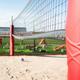 Beach volleyball court with a volleyball on the sand, visible net, and a red padded post support.