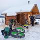 A man works on his skis next to parked bicycles at a log cabin.