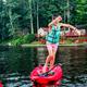 A young boy wearing a turquoise life vest and pink shorts balancing on a red paddleboard on the river, with trees and cabins in the background.