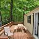 Wooden deck beside a beige cabin, with picnic table, chair, and wooded forest view.