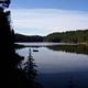 Glassy lake reflecting conifer lined shore, blue sky and cloud.