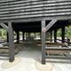 Eight picnic tables sit on a concrete pad under a wooden open sided picnic shelter