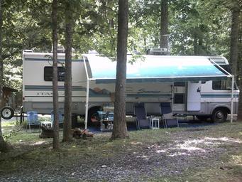 RV parked at a shaded campsite with awning, chairs and campsite setup — directly illustrates RV pitching and wooded campsite environment on the property.