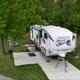 Concrete RV pad with a fifth-wheel parked, picnic table, grill and hookups visible — good view of RV sites and site amenities on the campground grounds.