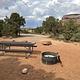 A campsite with metal picnic table and fire ring surrounded by pinyon and juniper trees and tall sandstone cliffs in the background.