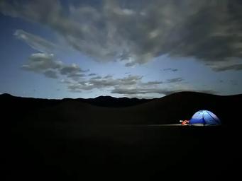 Nighttime. Glowing tent on dark sand dunes. A few stars and clouds in the sky.