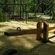 Wood chip covered play ground with wooden see saw and monkey bars next to tree lined roadway.