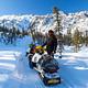 A man stands next to two snowmobiles in front of a rocky ridge.