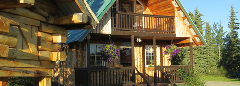 Exterior photo of a cabin with porch, hanging baskets and surrounding trees — from the site header/home page, shows cabin lodging appearance.