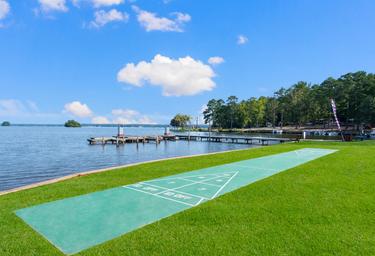 Lakeside view showing shuffleboard court, docks and shoreline — highlights waterfront amenities and open lawn. Image is part of the property photo set on the resort page (page: Lake Gaston RV Camping | Thousand Trails RV Campground, URL provided).