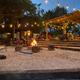 Outdoor gathering around a fire pit with seating. String lights illuminate a wooden structure and people.