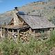 Log cabin set in brush, a full woodshed overlooking a hillside blanketed with silvered conifer snags.