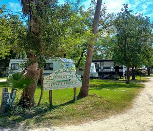 Bayou Campground -A weathered sign reading “Welcome, Office & Store, Private Property” stands near RVs and trees along a dirt road under a sunny sky.