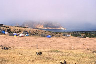 Tents and 5-foot tall windbreaks perches on an ocean bluff overlooking an islet covered in fog.