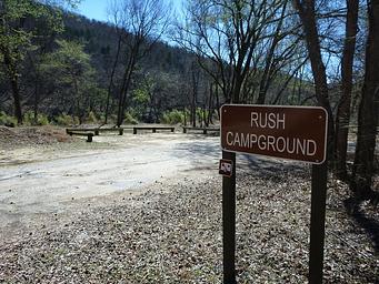 A brown sign stating "Rush Campground" can be seen in the foreground with a dirt road, trees, and a parking area in the background.
