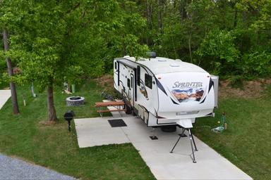Concrete RV pad with a fifth-wheel parked, picnic table, grill and hookups visible — good view of RV sites and site amenities on the campground grounds.