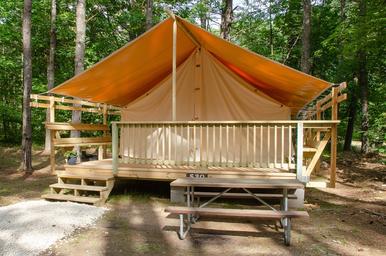 Front view of a raised canvas glamping/tent cabin with deck and picnic table in a wooded setting — matches the KOA ‘glamping’/cabin accommodations shown on the campground’s ways-to-stay photos page.