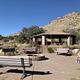 Uncovered fire pit surrounded by benches, with the covered pavilion with picnic table in the background.