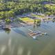 Aerial view of Lazy L Lake Campground showing RV sites, the sandy swimming beach lined with kayaks and floats, and the inflatable aqua park on the 75-acre lake