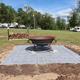 Ground-level view of a fire pit area with stacked wood and RVs parked in the background — highlights on-site amenity and campsite layout (gallery page).