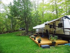 Ground-level view of an RV with a small deck on a grassy wooded site — illustrates seasonal RV campsite accommodations on the property (matches campsite references on the site).