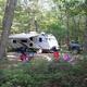 Daytime view of an RV parked in a wooded campsite with chairs and picnic setup — useful for showing campsite setting and surroundings (filename on site: ‘100 Sites’ image preview).