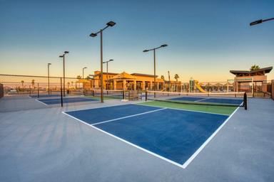 Empty outdoor pickleball courts with blue and green surfaces, surrounded by fencing and tall lights, near a modern building and a playground under a clear sky at sunset.