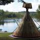 Tipi tent on a lake with people on the beach and boats.