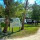 Bayou Campground -A weathered sign reading “Welcome, Office & Store, Private Property” stands near RVs and trees along a dirt road under a sunny sky.