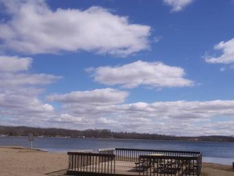 Sandy beach and lakeside deck with picnic tables looking across the lake — shows on-site waterfront recreation area referenced on the property page.