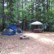 A campsite in a forest with a blue tent, a white canopy, and a picnic table.