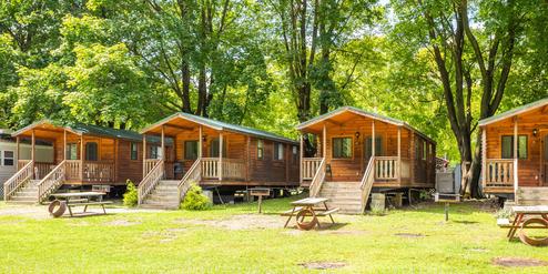 Row of wooden cabins with picnic tables on grassy, shaded lots — directly matches the site’s accommodations imagery on the Amenities/Accommodations pages.